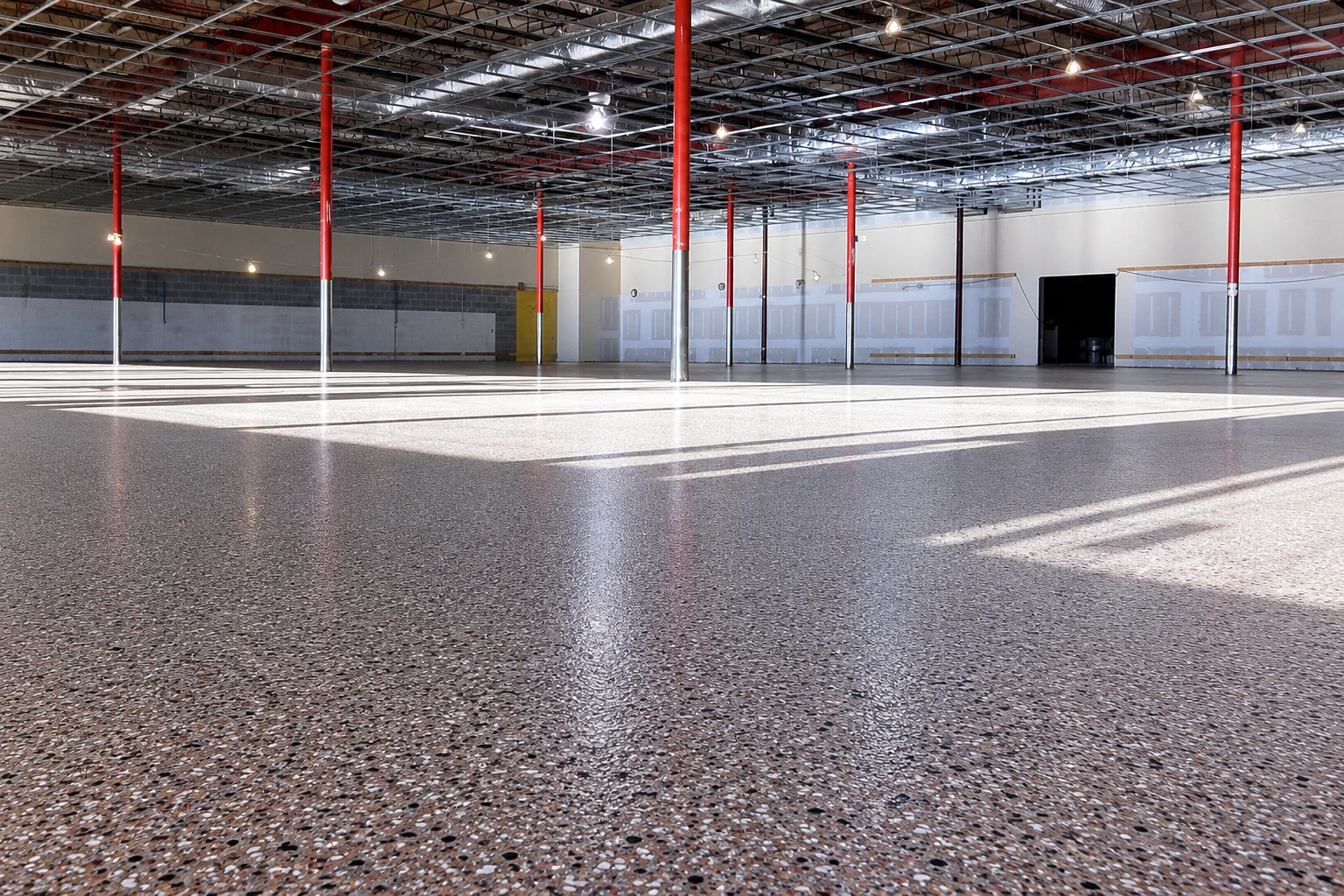 A polished speckled concrete floor spans a large unfinished commercial interior beneath exposed ceiling framing and red support columns.