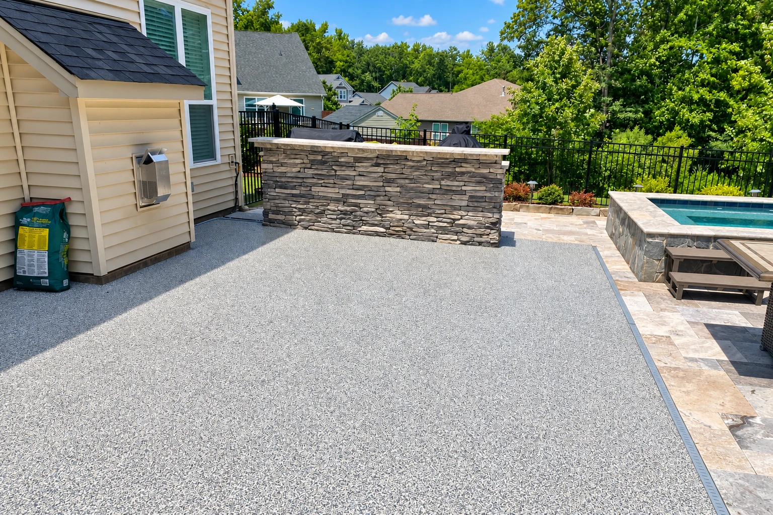 A broad textured gray concrete patio beside a house, with a stone outdoor counter and a swimming pool at the edge.