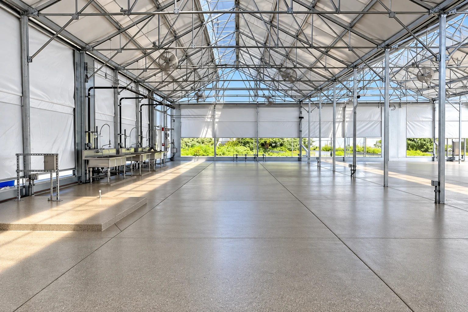 A smooth concrete floor with expansion joints fills a bright metal-framed indoor workspace lined with utility sinks.