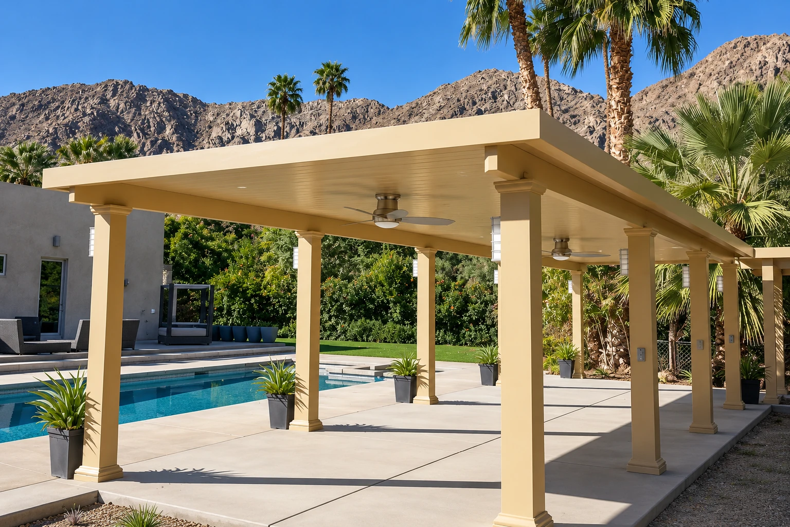 Concrete pool deck beneath a long cream-colored pergola, with a swimming pool, potted plants, palms, and rocky hills.
