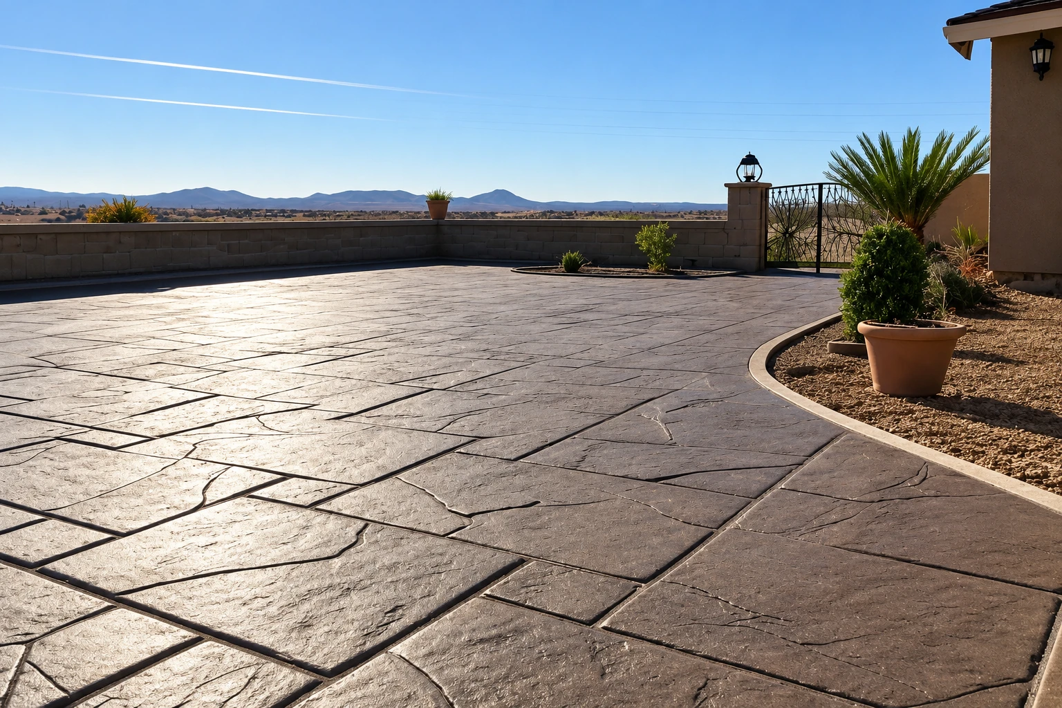 Stamped concrete patio with a dark gray stone pattern curves around a planted bed beside a low block wall and desert hills.
